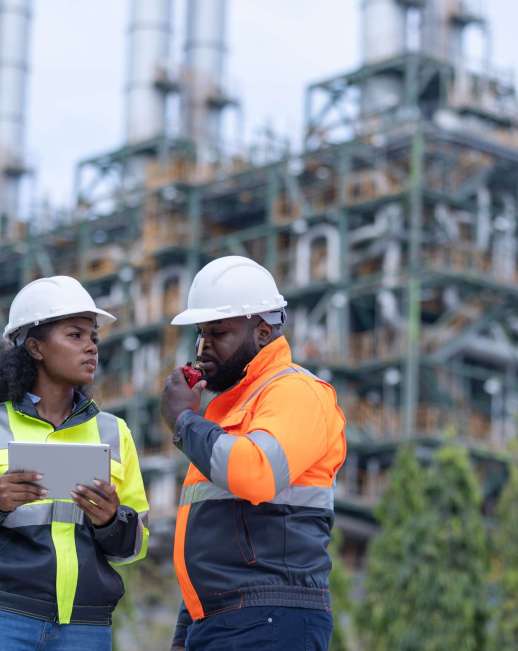 Two engineers wearing safety helmets and reflective jackets stand in front of a power plant, discussing plans while using a tablet and a radio during a site inspection.